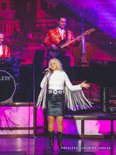 Woman with curly blonde hair who is wearing a white fringe shirt and leather skirt while performing a classic country song on a live show stage in Branson.