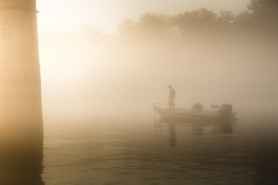 A boater stands on the bow of a small boat surrounded by morning mist on Lake Taneycomo