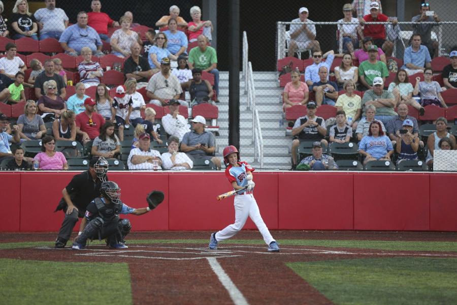 Youth baseball player hitting ball with people in the stands behind him.