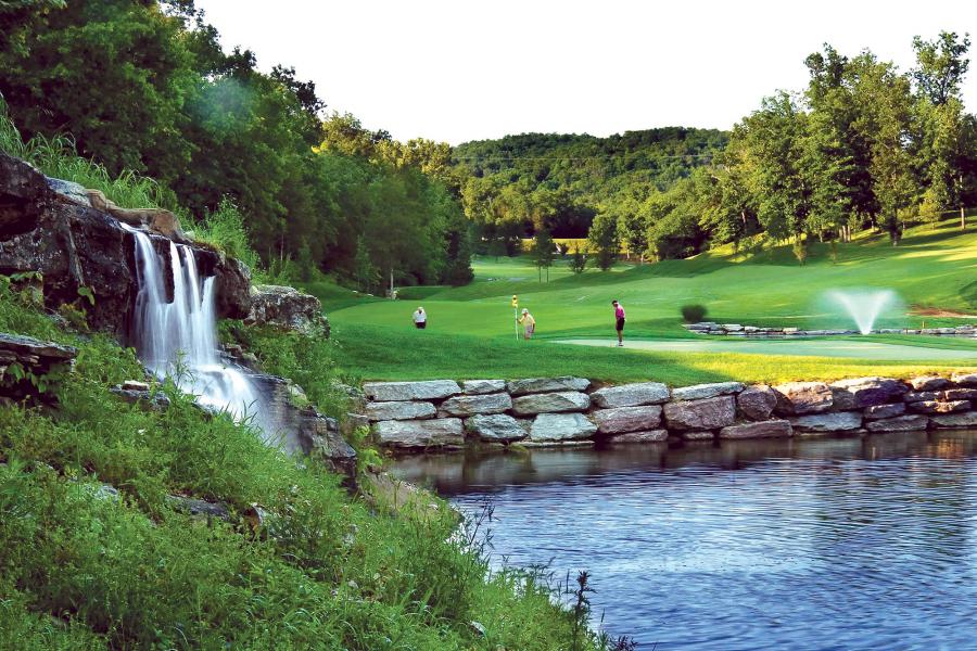 Group of golfers putting on a green with a small waterfall and pond in the foreground.