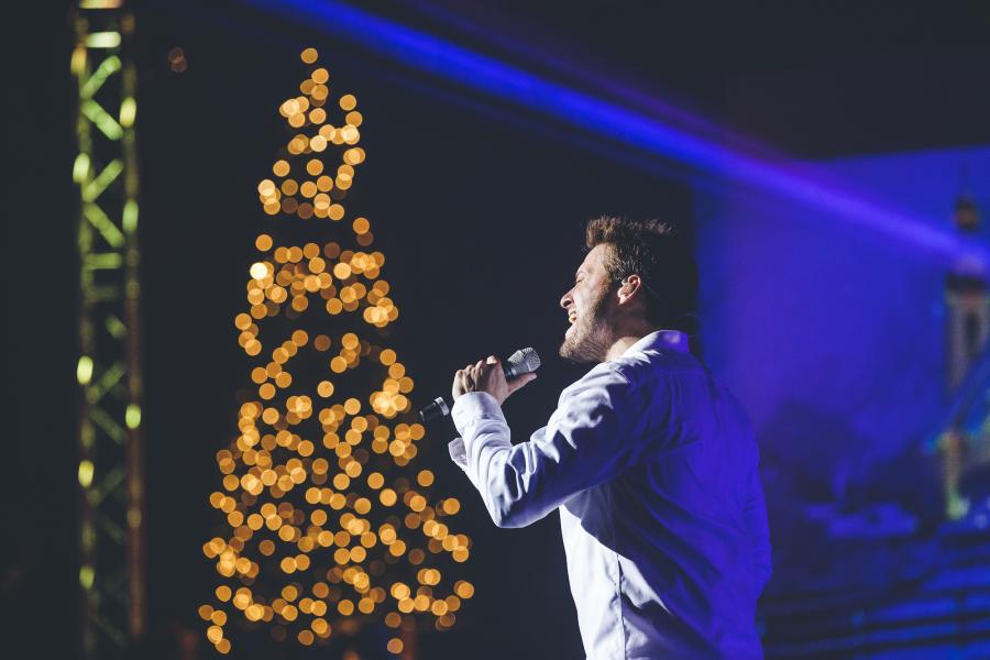 Singer from The Haygoods show singing into a microphone in front of a Christmas tree with lights.