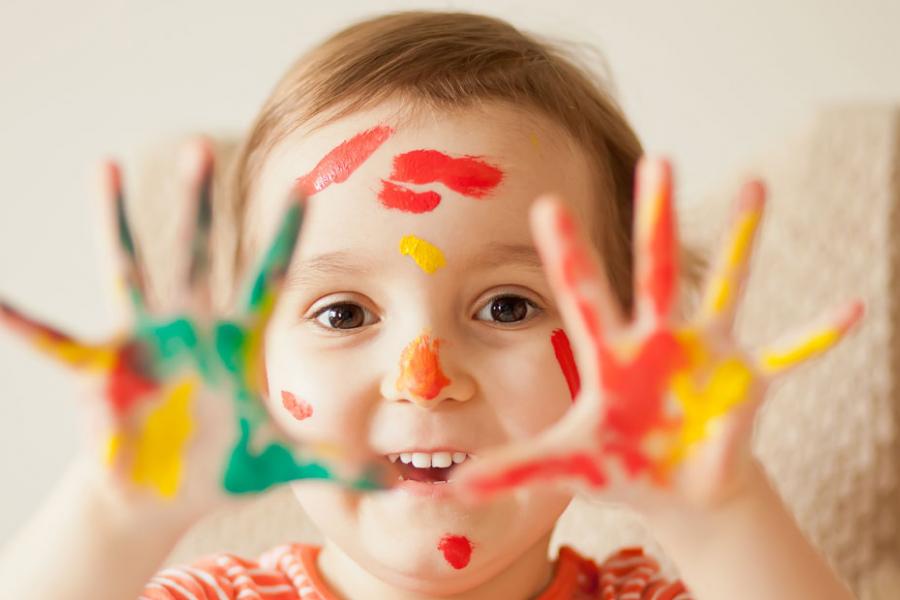 Young girl in striped orange shirt holding up her hands with bright paint splotches all over them.