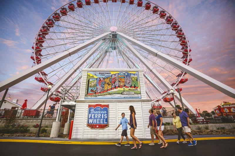 Family walks past a large ferris wheel and Branson mural at dusk.