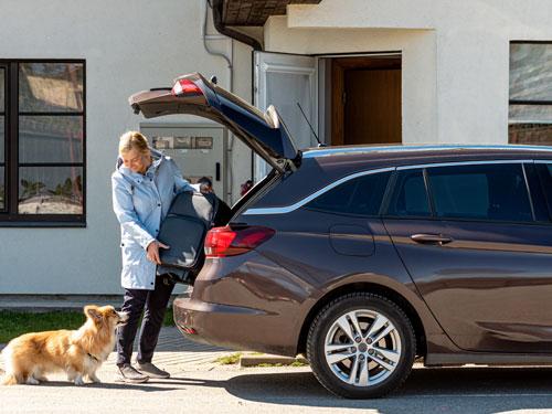 Woman wearing blue raincoat putting suitcase inside a brown car and looking at a dog.