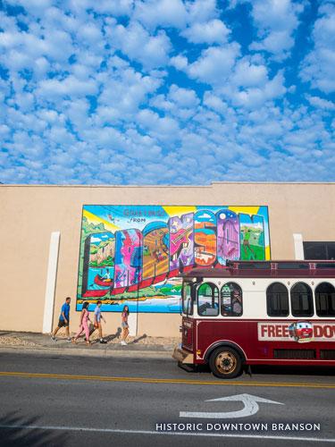 Family of four walking in front of Branson mural while free downtown trolley passes.