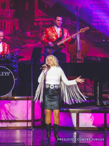 Woman with curly blonde hair who is wearing a white fringe shirt and leather skirt while performing a classic country song on a live show stage in Branson.