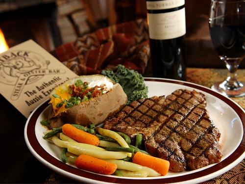 A plate of cooked vegetables, a loaded baked potato and a steak served with wine and an outback steakhouse menu.