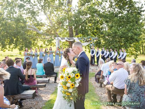 Recently married couple kissing at the end of the aisle at an outdoor wedding venue underneath a large sycamore tree.