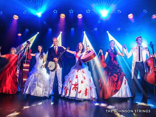 Family band performing on well-lit stage with violins, guitars and banjos.