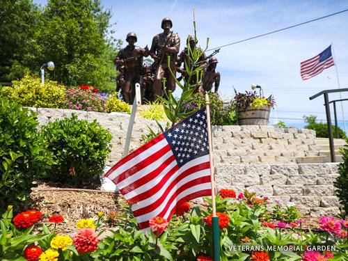 American flag in the foreground and statue of american soldiers in the background. The statue is surrounded by stunning flower displays in an outdoor garden.