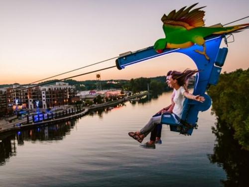 A man and women flying over a lake in Branson on a zipline towards an outdoor shopping center.