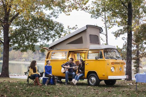 A family sits outside, lakeside next to their Volkswagen pop-top camper van and dad plays the guitar.