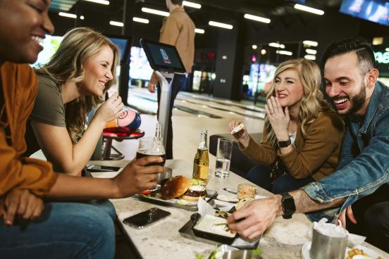 Group of friends eating and drinking around table with bowling lanes in the background at Andy B's Social in Branson.