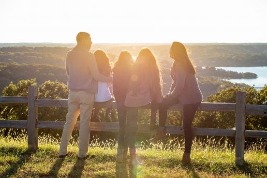 Family sits on fence while enjoying sunset over rolling hills and a lake.
