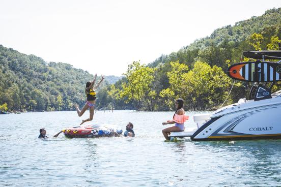 Young girl jumps off boating tube into a lake while mom watches on the back of the boat.