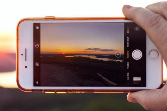 Hand holding a phone that is taking pictures of a scenic sunset and distant lake.