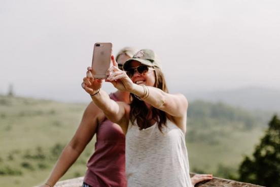 Two women surrounded by a beautiful nature scene are taking a selfie with their phone camera.