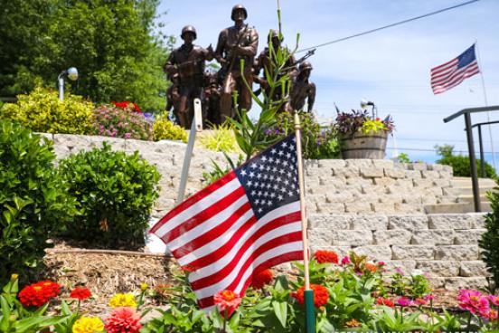 American flag in the foreground and statue of american soldiers in the background. The statue is surrounded by stunning flower displays in an outdoor garden.