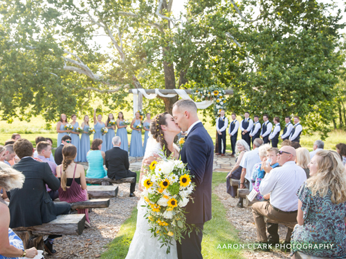 Recently married couple kissing at the end of the aisle at an outdoor wedding venue underneath a large sycamore tree.